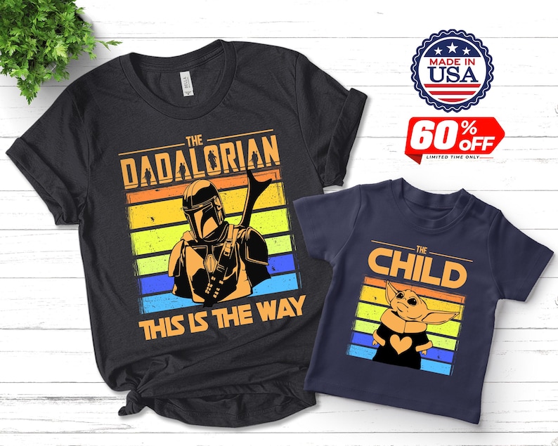 May include: Two t-shirts, one black and one navy blue, featuring graphics from a popular science fiction series. The black shirt says "The Dadalorian" with an illustration. The navy shirt says "The Child" with a character illustration. Both shirts have a retro sunset design.