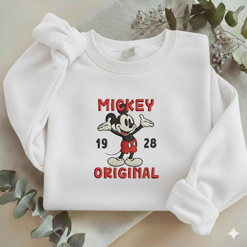 May include: A white long-sleeve sweatshirt featuring an embroidered graphic of Mickey Mouse in the center. The text above reads MICKEY and below reads ORIGINAL in red letters, with the numbers 19 and 28 positioned on either side of the character.