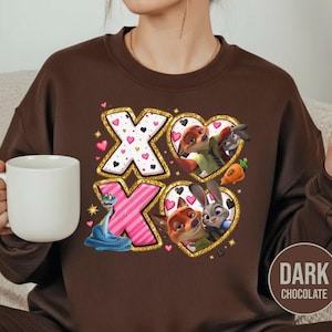 May include: Dark chocolate brown sweatshirt featuring a graphic design with the letters "XO" in gold glitter, pink, and white. The design includes cartoon characters in heart-shaped frames, with a blue snake and pink hearts.