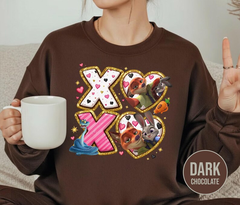 May include: Dark chocolate brown sweatshirt featuring a graphic design with the letters "XO" in gold glitter, pink, and white. The design includes cartoon characters in heart-shaped frames, with a blue snake and pink hearts.
