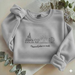 May include: A light grey crewneck sweatshirt featuring a black line art graphic of a castle, a mouse ear icon, a tree, and a geodesic sphere. Below the illustrations, the text reads "Happiest place on earth" in a cursive script font.