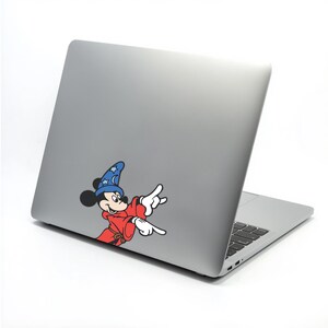 May include: A silver laptop with a sticker of Mickey Mouse dressed as a wizard. Mickey is wearing a blue hat with stars, a red robe, and white gloves, pointing with both hands. The laptop is on a white background.