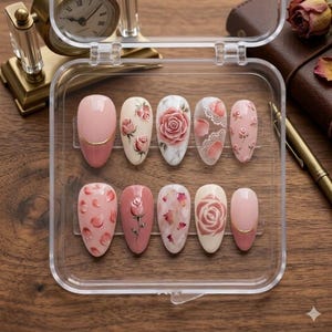 May include: A set of ten artificial nails in a clear plastic case. The nails feature various designs, including roses, floral patterns, and solid colors. Some nails have a gold accent. The color palette includes shades of pink, white, and beige.