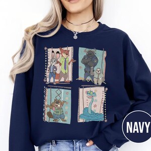 K&ouml;nnte beinhalten: Marineblaues Sweatshirt mit vier gerahmten Illustrationen von Zootopia-Charakteren. Die Illustrationen zeigen Judy Hopps, Nick Wilde, Chief Bogo, Flash, Mr. Big und eine Schlange. Das Wort "NAVY" ist in einem wei&szlig;en Kreis unten rechts aufgedruckt.