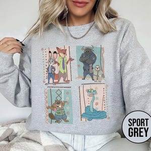 K&ouml;nnte beinhalten: Sportgraues Sweatshirt mit einem farbenfrohen Grafikdesign von Zootopia-Charakteren. Das Design umfasst Judy Hopps, Nick Wilde, Chief Bogo, Flash und andere. Das Sweatshirt hat einen Rundhalsausschnitt und lange &Auml;rmel.