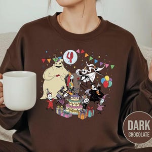 May include: Dark chocolate brown sweatshirt featuring a colorful graphic of characters from The Nightmare Before Christmas celebrating a birthday. The design includes a cake, balloons, and the number 4. The sweatshirt is a crew neck style.