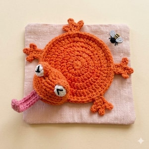 May include: A handmade orange crochet frog with white eyes and a pink tongue, lying on a pink square. A small embroidered bee is near the frog. The frog has four orange feet and is made of yarn.