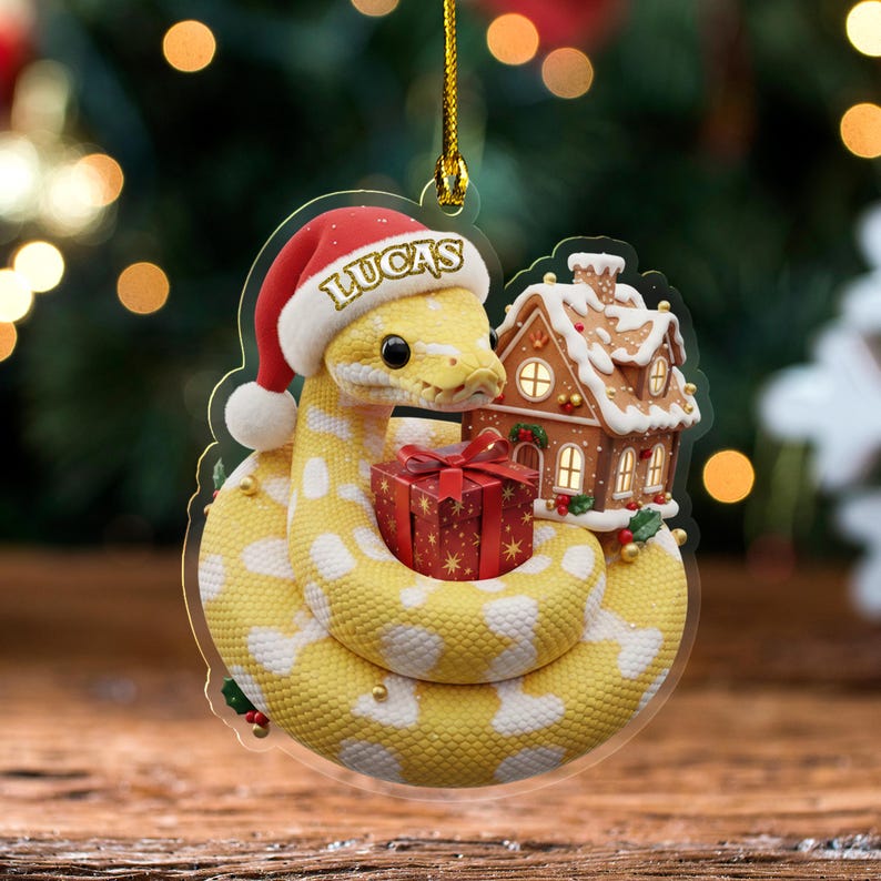 May include: A Christmas tree decoration showing a yellow snake wearing a Santa hat with the name "Lucas". The snake is coiled around a red gift box and a gingerbread house. The ornament has a gold hanging string.