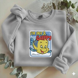 May include: A light gray crewneck sweatshirt featuring a central embroidered graphic of a yellow fish with blue stripes. The text above the fish reads I'M NOT A GUPPY in red and yellow, with the word Flounder written in cursive below the fish.
