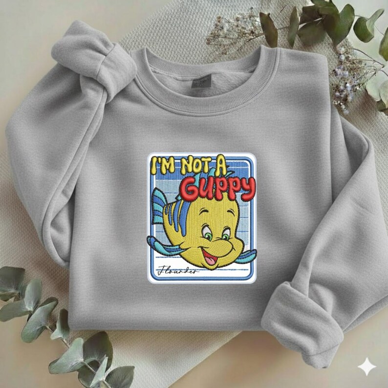 May include: A light gray crewneck sweatshirt featuring a central embroidered graphic of a yellow fish with blue stripes. The text above the fish reads I'M NOT A GUPPY in red and yellow, with the word Flounder written in cursive below the fish.
