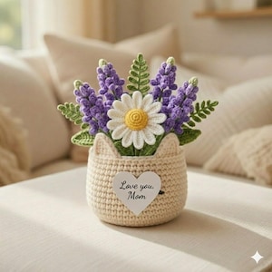 May include: A crocheted flower arrangement in a cat-shaped basket. The arrangement includes purple lavender, a white daisy with a yellow center, and green leaves. The basket is beige with a heart-shaped tag that reads "Love you, Mom."