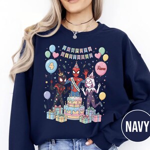 May include: Navy blue sweatshirt featuring a colorful graphic of Spider-Man characters celebrating a birthday. The design includes a cake, balloons, and the text "HAPPY BIRTHDAY" and "4". The word "Name" is on a balloon. The sweatshirt is a comfortable, casual style.