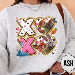 May include: A light gray sweatshirt featuring a design with the letters "XO" in gold and pink, with heart accents. The design includes cartoon characters in heart-shaped frames. The sweatshirt has long sleeves.