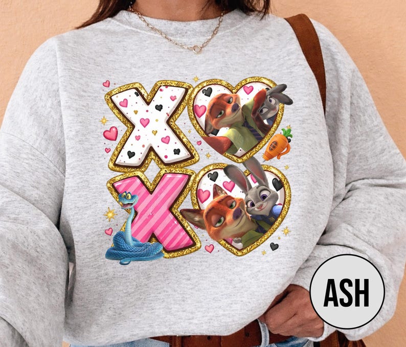 May include: A light gray sweatshirt featuring a design with the letters "XO" in gold and pink, with heart accents. The design includes cartoon characters in heart-shaped frames. The sweatshirt has long sleeves.