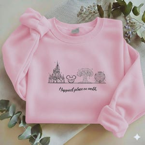 May include: A light pink crewneck sweatshirt featuring a black line art graphic of a castle, a mouse ear icon, a tree, and a geodesic dome. Below the illustrations, the text reads, "Happiest place on earth" in a black cursive script font.