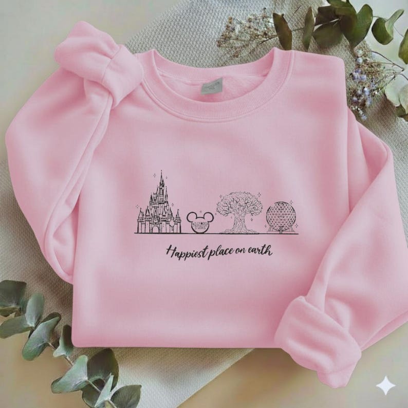 May include: A light pink crewneck sweatshirt featuring a black line art graphic of a castle, a mouse ear icon, a tree, and a geodesic dome. Below the illustrations, the text reads, "Happiest place on earth" in a black cursive script font.