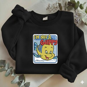 May include: A black crewneck sweatshirt featuring a central embroidered graphic of a yellow and blue fish character. The text reads I'M NOT A GUPPY in bold red and yellow letters, with the name Flounder written in cursive script below the fish.