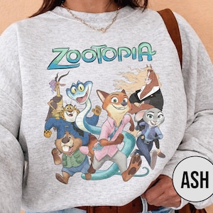 以下が含まれることがあります： 水色の「Zootopia」の文字と、アニメ映画のキャラクターのカラフルなグラフィックが特徴のライトグレーのスウェットシャツ。デザインには、キツネ、ウサギ、ヘビ、その他の動物のキャラクターが含まれています。