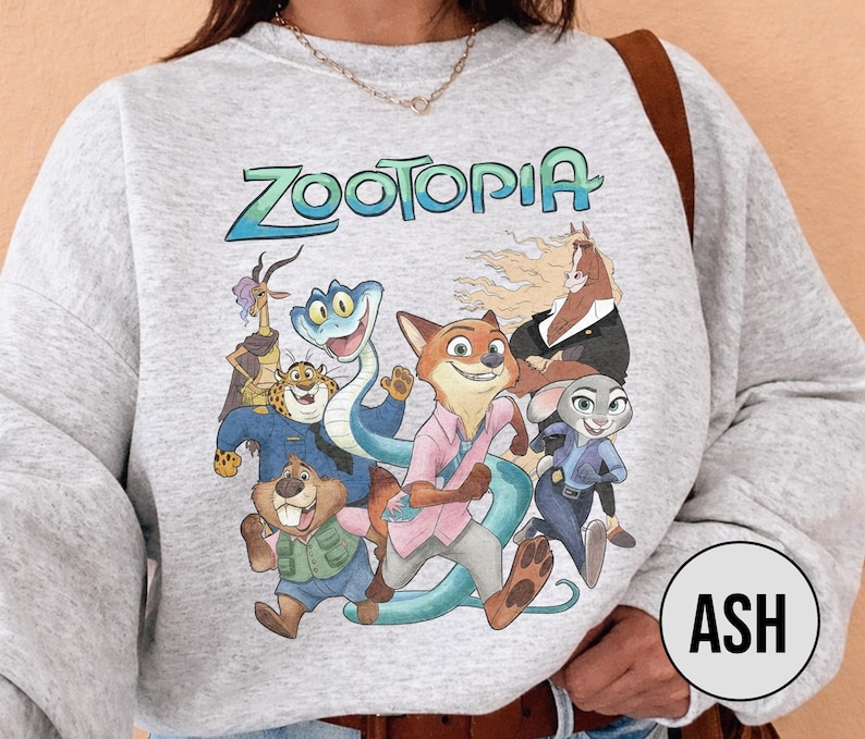 以下が含まれることがあります： 水色の「Zootopia」の文字と、アニメ映画のキャラクターのカラフルなグラフィックが特徴のライトグレーのスウェットシャツ。デザインには、キツネ、ウサギ、ヘビ、その他の動物のキャラクターが含まれています。