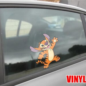 May include: Orange and tan vinyl decal of a cartoon character with large ears and a tiger-like pattern, waving with a cheerful expression. The decal is applied to a car window. The word "VINYL" is printed in red.