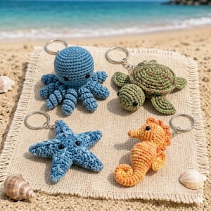 May include: Four crochet keychains in the shape of sea creatures: a blue octopus, a green turtle, a blue starfish, and an orange seahorse. Each keychain has a silver ring. The keychains are on a beige cloth, with a beach and ocean in the background.