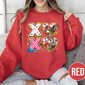 May include: A red sweatshirt featuring the text "XO" with heart and character graphics. The design includes cartoon characters within heart shapes, and a blue snake. The sweatshirt has a round neckline and long sleeves.