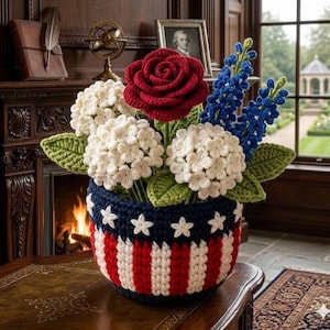 May include: A handcrafted crochet floral arrangement in a patriotic theme. The arrangement features a red rose, white hydrangeas, and blue flowers, set in a basket with red and white stripes and white stars on a navy blue background.