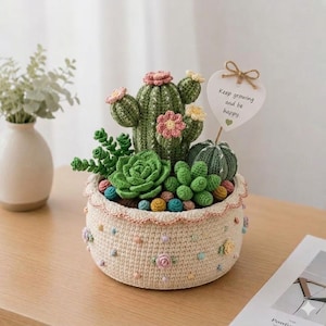 May include: A crocheted cactus and succulent arrangement in a beige pot. The arrangement includes green cacti, succulents, and colorful decorative balls. A heart-shaped sign with the text "Keep growing and be happy" is attached.
