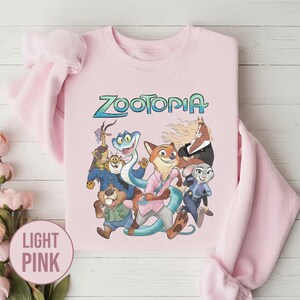 以下が含まれることがあります： ターコイズ色の「Zootopia」の文字と、アニメ映画のキャラクターのグラフィックが特徴の、薄いピンク色のスウェットシャツ。長袖のトップスは丸首です。「Light Pink」の文字が左下に円で表示されています。