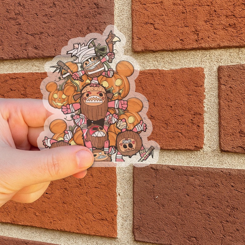 May include: A clear sticker featuring a group of cartoon characters with pumpkin heads and wrapped in bandages. The characters are in shades of brown, orange, and pink, with white accents. The sticker is held against a red brick wall.