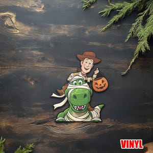 May include: A Halloween-themed vinyl sticker featuring Woody from Toy Story riding a green dinosaur wrapped in bandages. Woody holds a jack-o'-lantern. The word "VINYL" is in a red box at the bottom.