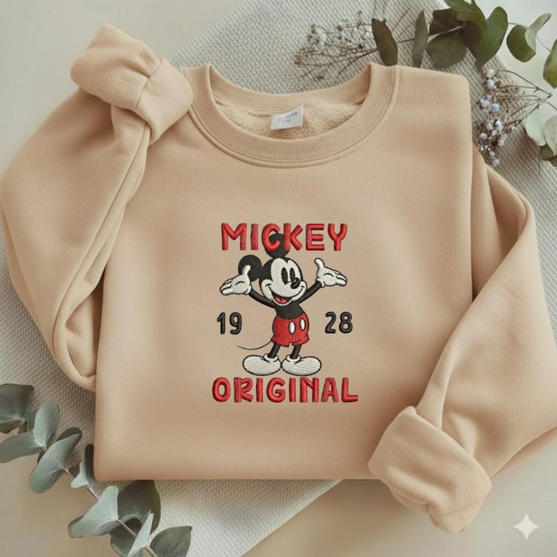 May include: A beige crewneck sweatshirt featuring an embroidered graphic of Mickey Mouse in the center. The text reads MICKEY ORIGINAL with the numbers 19 and 28 on either side of the character. The design uses red, black, and white thread colors.