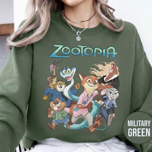 以下が含まれることがあります： 「Zootopia」の文字とアニメ映画のキャラクターのグラフィックが特徴のミリタリーグリーンのスウェットシャツ。デザインにはキツネ、ウサギ、ヘビ、その他の動物のキャラクターが含まれています。スウェットシャツは無地です。