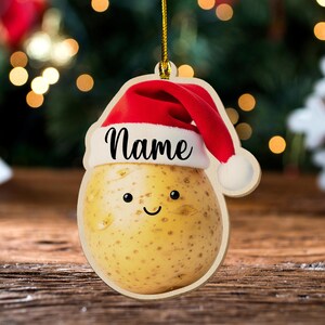 May include: A wooden Christmas ornament shaped like a potato wearing a Santa hat. The ornament has the word "Name" in black script and a smiling face. The potato is light brown with small spots.