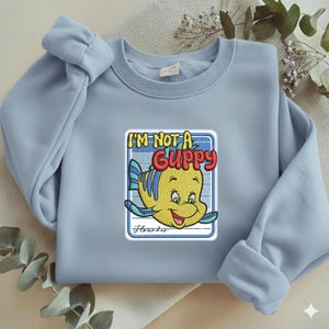 May include: A light blue crewneck sweatshirt featuring a central embroidered patch of a yellow and blue fish character. The text above the fish reads I'M NOT A GUPPY in bold red letters, and the name Flounder is written in cursive below the fish.