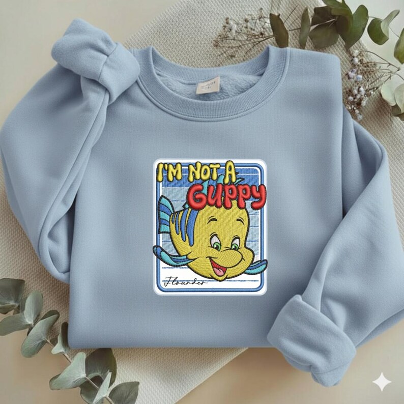 May include: A light blue crewneck sweatshirt featuring a central embroidered patch of a yellow and blue fish character. The text above the fish reads I'M NOT A GUPPY in bold red letters, and the name Flounder is written in cursive below the fish.