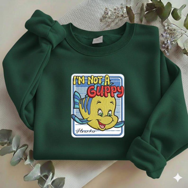 May include: A dark green crewneck sweatshirt featuring a central embroidered patch of a yellow fish with blue fins. The text reads I'M NOT A GUPPY in red and yellow letters, with the name Flounder written in cursive script below the fish illustration.