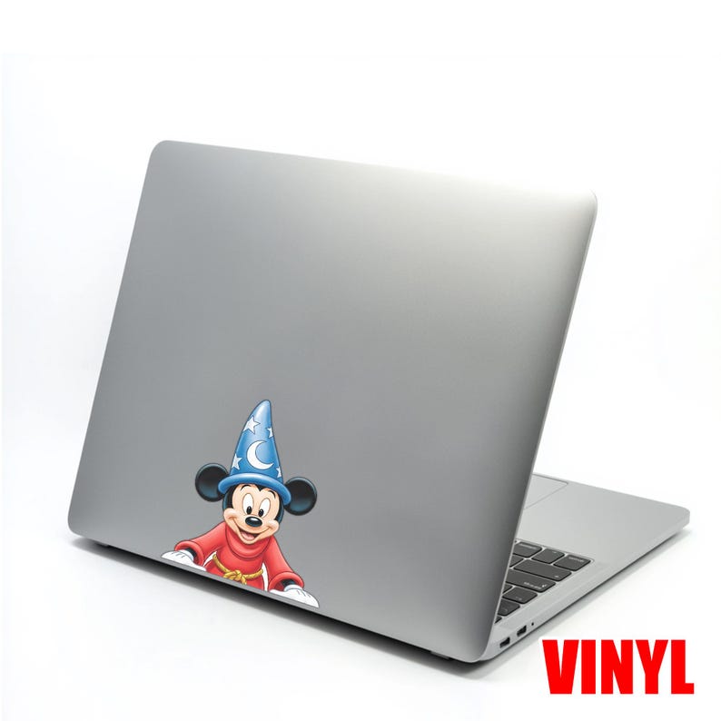 May include: A silver laptop with a vinyl sticker of Mickey Mouse in a blue wizard hat and red robe. The sticker is on the back of the laptop. The word "VINYL" is printed in red on the bottom right corner.