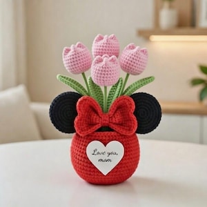 May include: A handmade crochet flower arrangement featuring pink tulips, green leaves, and a red vase with a bow and black ears. The vase has a white heart with the text "Love you, mom."