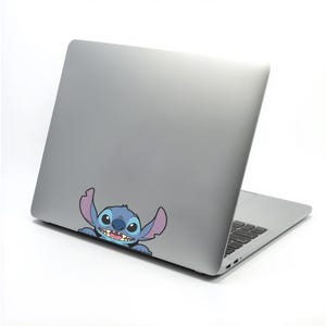 May include: A silver laptop with a cartoon sticker of Stitch, a blue alien character with large ears and a toothy grin. The sticker is placed on the lower left corner of the laptop's lid.