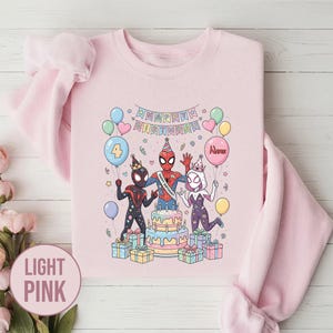 May include: Light pink sweatshirt featuring a colorful illustration of three Spider-Man characters celebrating a birthday. The design includes a cake, balloons, and the words "Happy Birthday". The sweatshirt is a crew neck style.
