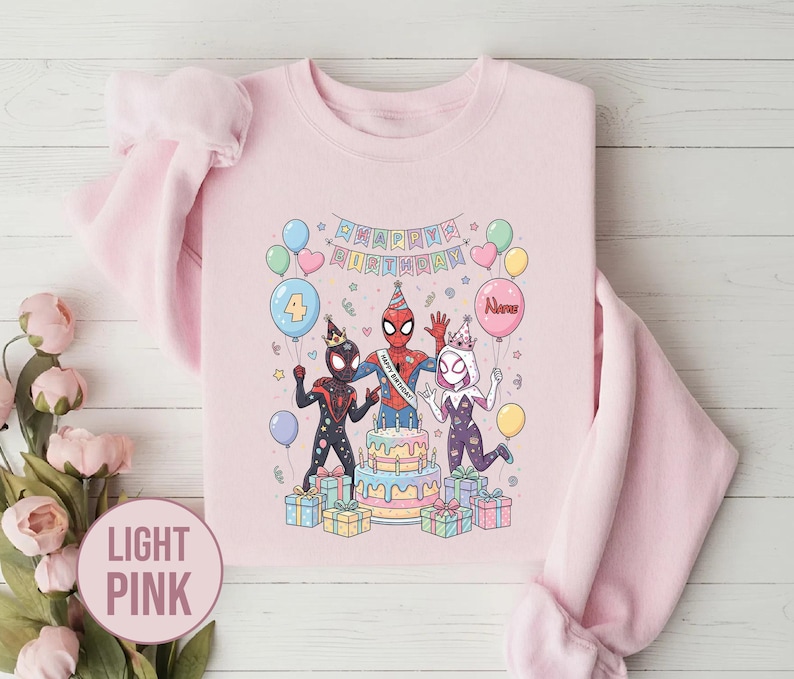 May include: Light pink sweatshirt featuring a colorful illustration of three Spider-Man characters celebrating a birthday. The design includes a cake, balloons, and the words "Happy Birthday". The sweatshirt is a crew neck style.