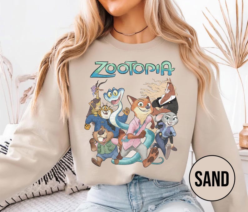 以下が含まれることがあります： 「Zootopia」の文字が青と緑で描かれたサンドカラーのスウェットシャツ。アニメ映画のキャラクターのグラフィックが特徴です。デザインには、キツネ、ウサギ、ヘビ、その他の動物のキャラクターが含まれています。