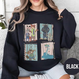 K&ouml;nnte beinhalten: Schwarzes Sweatshirt mit einem farbenfrohen Grafikdesign von Charakteren aus dem Animationsfilm Zoomania. Das Design umfasst Judy Hopps, Nick Wilde, Chief Bogo, Flash und andere. Das Wort "BLACK" ist auf einem schwarzen Kreis unten rechts aufgedruckt.