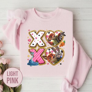 May include: Light pink sweatshirt with a graphic design featuring the characters from Zootopia. The design includes the letters "XO" with heart and character illustrations. The text "LIGHT PINK" is in a pink circle.