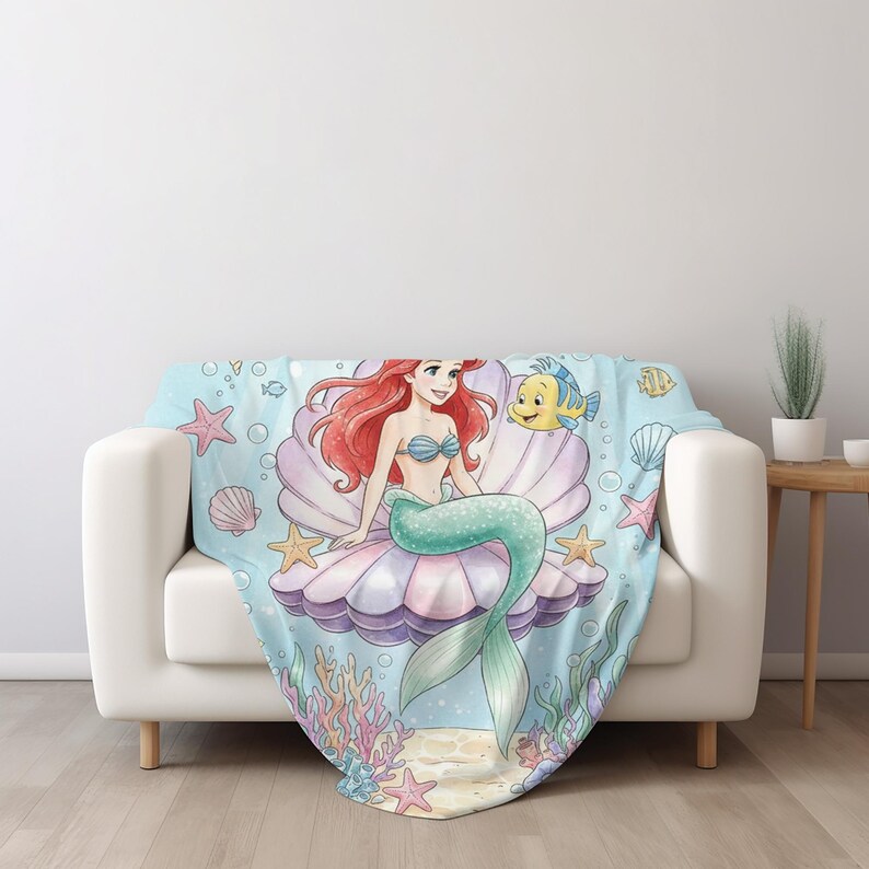 K&ouml;nnte beinhalten: Eine Decke mit einer Illustration einer Meerjungfrau mit roten Haaren und einem gr&uuml;nen Schwanz, die in einer Muschel sitzt. Die Decke hat einen hellblauen Hintergrund mit Seesternen und Blasen. Die Decke liegt auf einem Sofa.