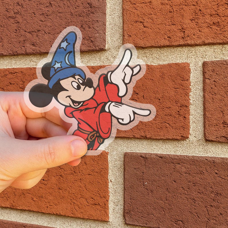 May include: A clear sticker featuring Mickey Mouse in a red robe and blue wizard hat with stars. Mickey is pointing with both hands. The sticker is held in front of a brick wall.