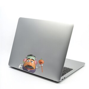 May include: A silver laptop with a sticker of a cartoon Mr. Potato Head dressed as Frankenstein, holding a pumpkin lollipop. The sticker has a green and black hat, a large mustache, and a purple vest. The pumpkin is orange.