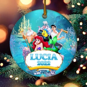 May include: A round Christmas ornament featuring characters from The Little Mermaid. The ornament has a teal background with the characters Ariel, Flounder, Sebastian, Ursula, Prince Eric, and King Triton. The name "Lucia" and the year "2022" are displayed.