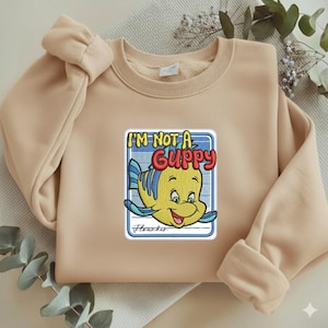 May include: A beige crewneck sweatshirt featuring a central embroidered patch of a yellow and blue fish character. The text reads I'M NOT A GUPPY in bold red letters above the fish, with the name Flounder written in cursive script below.